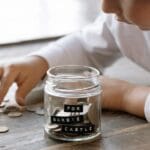 A young child collects coins in a jar labeled 'For Barbie Castle', symbolizing saving and dreams.