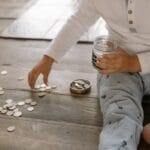 A young child collects coins into a jar on a wooden floor, symbolizing savings.