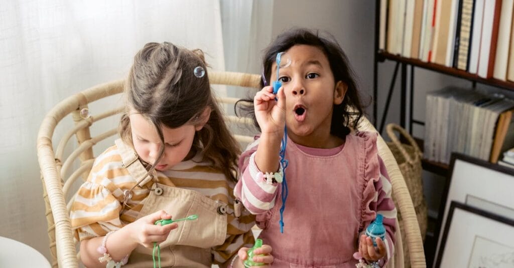 Two girls joyfully blowing bubbles indoors, surrounded by books.