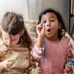 Two girls joyfully blowing bubbles indoors, surrounded by books.