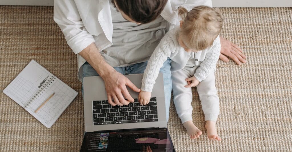 A father and baby sitting on a carpeted floor, interacting with a laptop in a cozy setting.