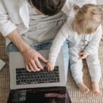 A father and baby sitting on a carpeted floor, interacting with a laptop in a cozy setting.