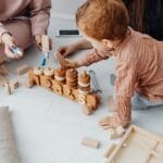 A young child engaged with a wooden toy train in a warm indoor setting, surrounded by adults.