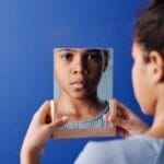 A young girl holds a mirror, reflecting her face, set against a vivid blue background.