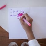 A child writes 'I love you mom' on a card with pink and purple pens on a white table.