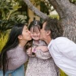 Happy parents kissing their daughter with Down syndrome in a loving outdoor setting.