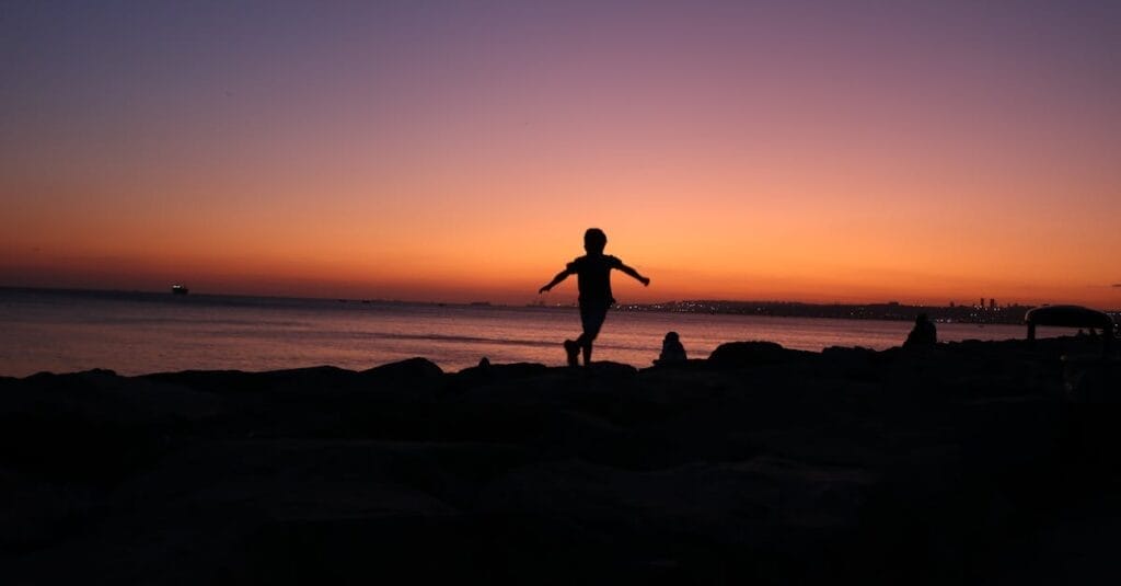 Back view of silhouette anonymous child running along sea shore against sunset sky with colorful gradient