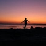 Back view of silhouette anonymous child running along sea shore against sunset sky with colorful gradient