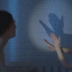 A mother and child creating shadow puppets on a bedroom wall under soft lighting.