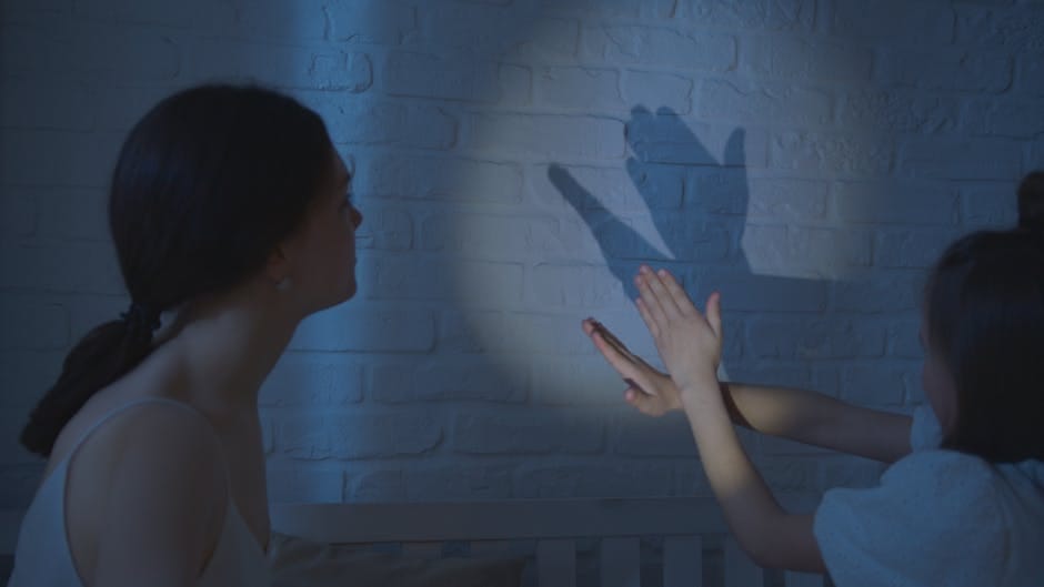 A mother and child creating shadow puppets on a bedroom wall under soft lighting.