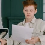 A young boy sits at a piano, reading sheet music, focused on learning indoors.