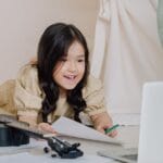 Adorable child engaging in online learning with a ukulele and laptop indoors.