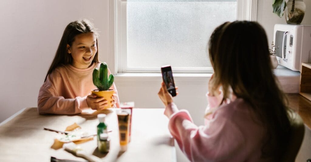 Two girls enjoy arts and crafts indoors, with one capturing the moment with a smartphone.