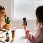 Two girls enjoy arts and crafts indoors, with one capturing the moment with a smartphone.