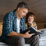 A father and son share a warm moment reading a book in a cozy attic bedroom.