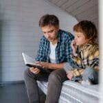 A father and son share a bonding moment while reading together in a cozy bedroom setting.