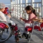 Two young girls enjoying a sunny day on a terrace, one in a wheelchair, sharing a sweet moment.