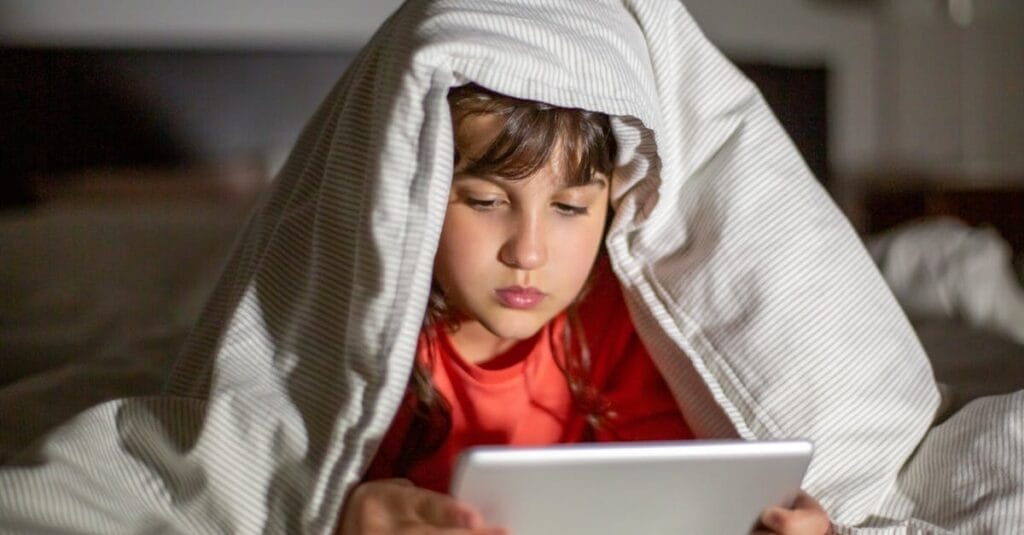 Child using a tablet under a blanket in bed, focused and engaged at night.