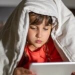 Child using a tablet under a blanket in bed, focused and engaged at night.