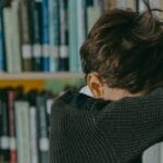 A young boy looking down, leaning against a bookshelf in a library, evoking a sense of loneliness.