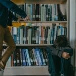 A child facing bullying while sitting in a library against bookshelves, depicting harassment and isolation.