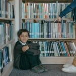 A sad boy sitting on the library floor while being bullied by a peer.