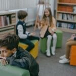 A sad child isolated in a school library while peers chat in the background.