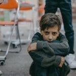 A young boy sits on the floor of a classroom, expressing fear and vulnerability.