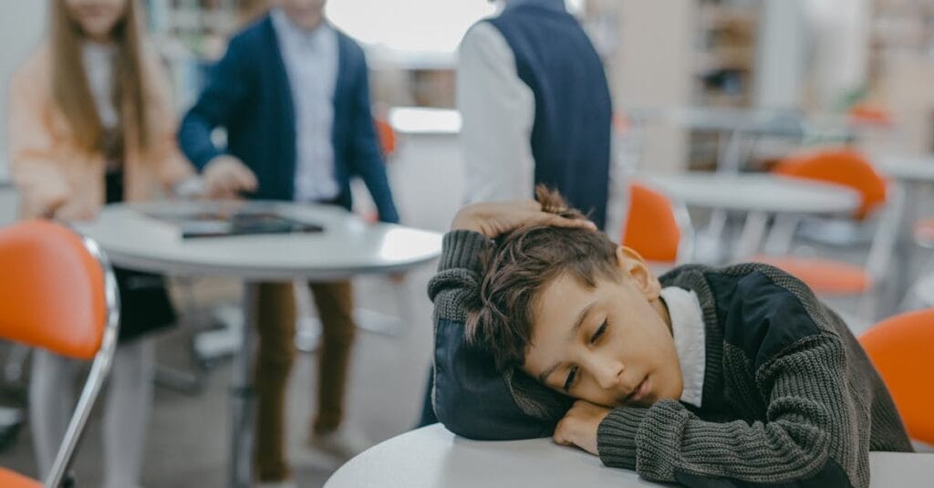 A sad child rests on a classroom desk while peers talk, symbolizing loneliness and school exclusion.