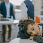 A sad child rests on a classroom desk while peers talk, symbolizing loneliness and school exclusion.