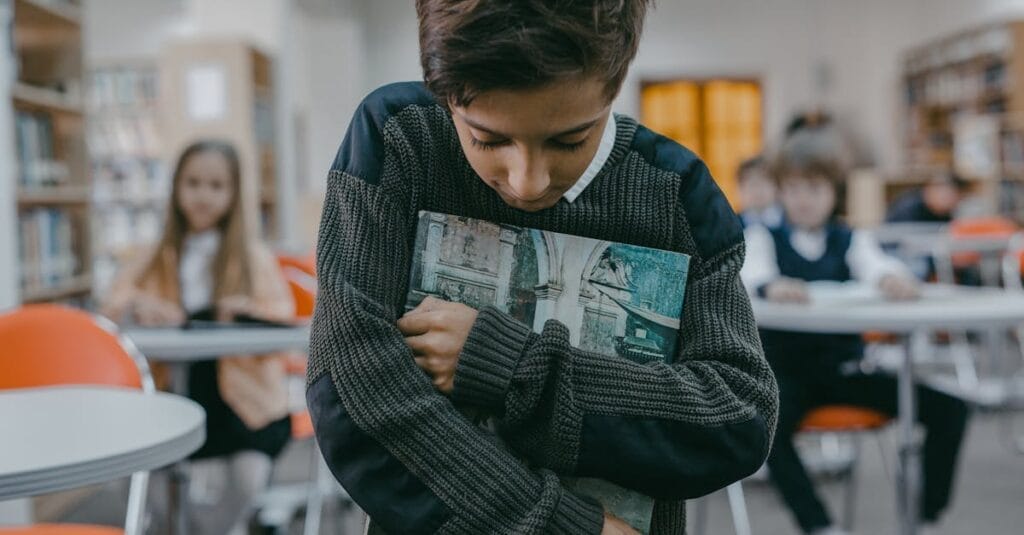 Boy stands alone, hugging book in a classroom, expressing sadness and isolation.