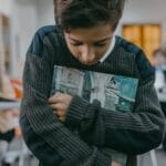 Boy stands alone, hugging book in a classroom, expressing sadness and isolation.