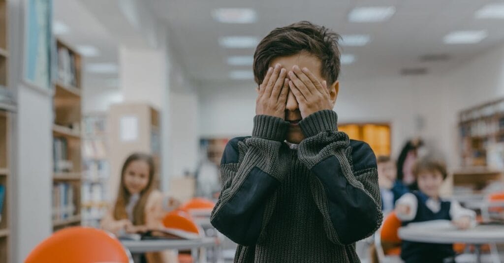 Young boy covers face in a bustling school classroom, indicating stress or anxiety.