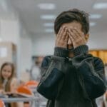 Young boy covers face in a bustling school classroom, indicating stress or anxiety.