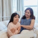 A mother and daughter share a joyful moment sitting on a bed in their cozy bedroom.