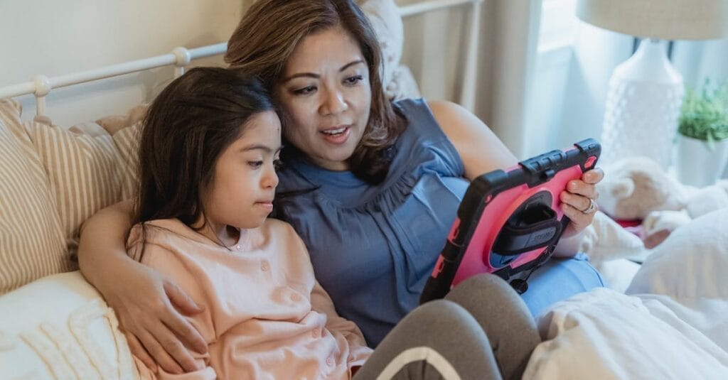 A mother and daughter share a moment, watching a tablet together in a cozy bedroom setting.