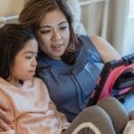 A mother and daughter share a moment, watching a tablet together in a cozy bedroom setting.