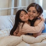 A mother and daughter happily embrace on a comfy bed, showcasing love and connection indoors.