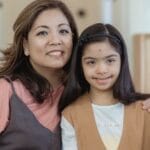 A mother and daughter with Down syndrome embracing and smiling indoors.