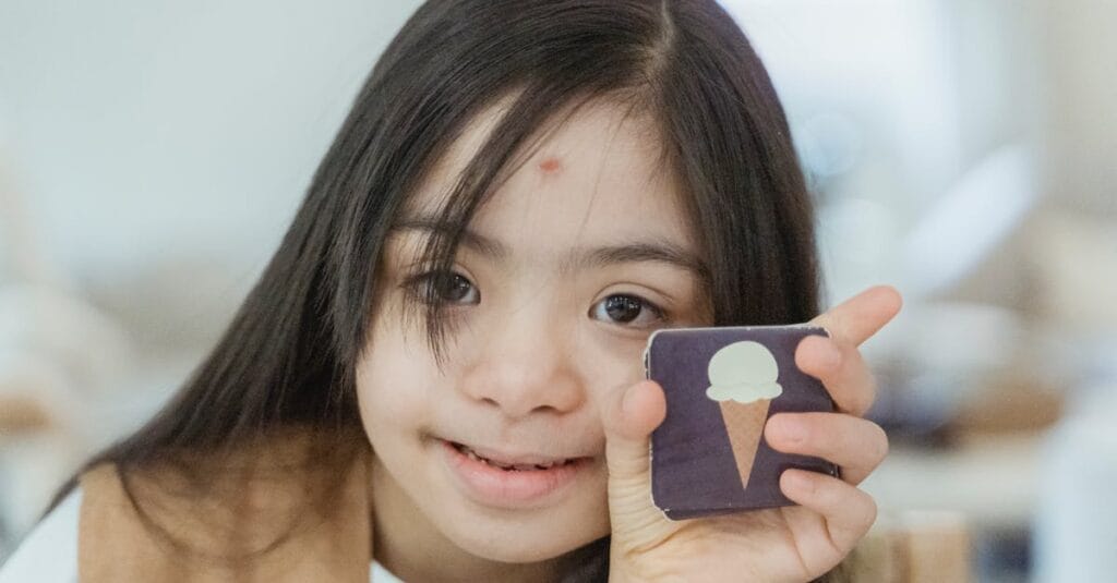 Young girl with Down syndrome smiling and holding a memory game card indoors.