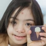 Young girl with Down syndrome smiling and holding a memory game card indoors.