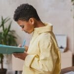 A young boy writing in a notebook while standing indoors, focused and engaged.