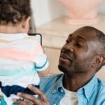 A father holding his young child indoors, sharing a joyful moment at home.