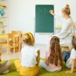 Teacher instructing young students with math on a chalkboard in a bright classroom setting.