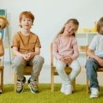 Group of children sitting on chairs, smiling inside a modern classroom setting.