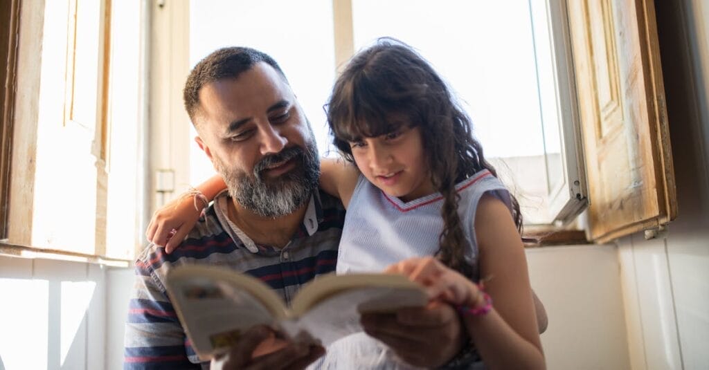 A loving father reads with his daughter by a sunlit window, fostering family bonding.