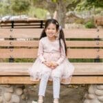 A joyful young girl in a pink dress sits smiling on a wooden bench in a serene park setting.