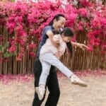 Father joyfully lifts daughter in a garden with vivid pink flowers and wooden fence.