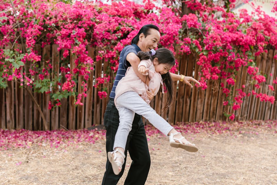 Father joyfully lifts daughter in a garden with vivid pink flowers and wooden fence.