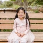 Cheerful young girl in a pink dress smiling while sitting on a park bench outdoors.